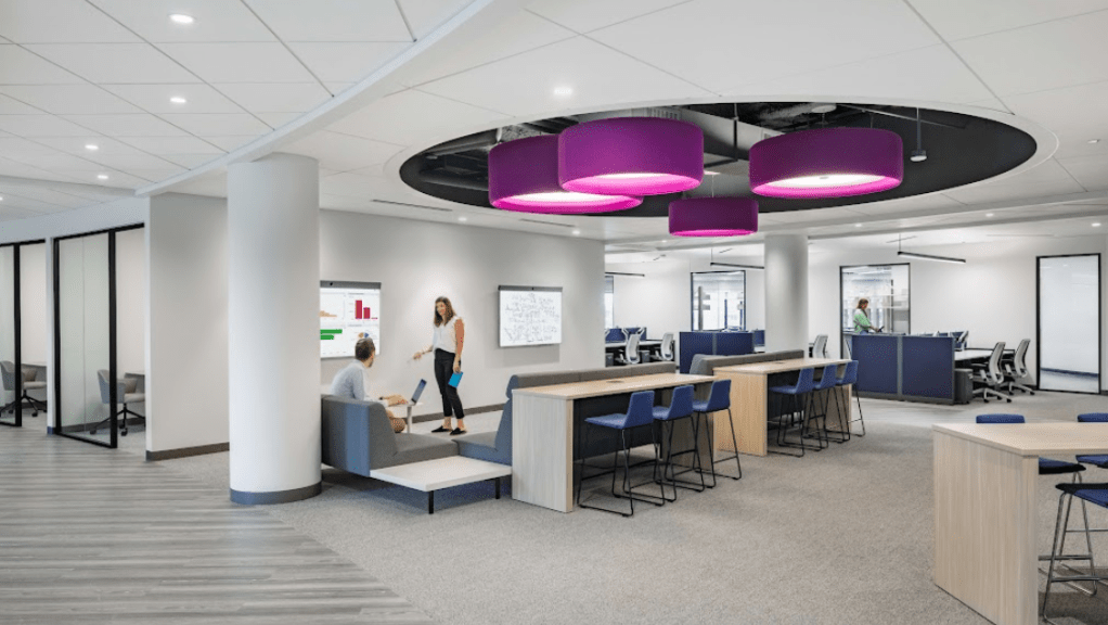 A modern open-plan office space with light-colored walls and furniture. In the foreground, a man and woman are collaborating near a screen displaying data charts. The room features high tables with barstools, private glass-enclosed meeting rooms, and bright purple circular light fixtures hanging from the ceiling, creating a dynamic atmosphere. 