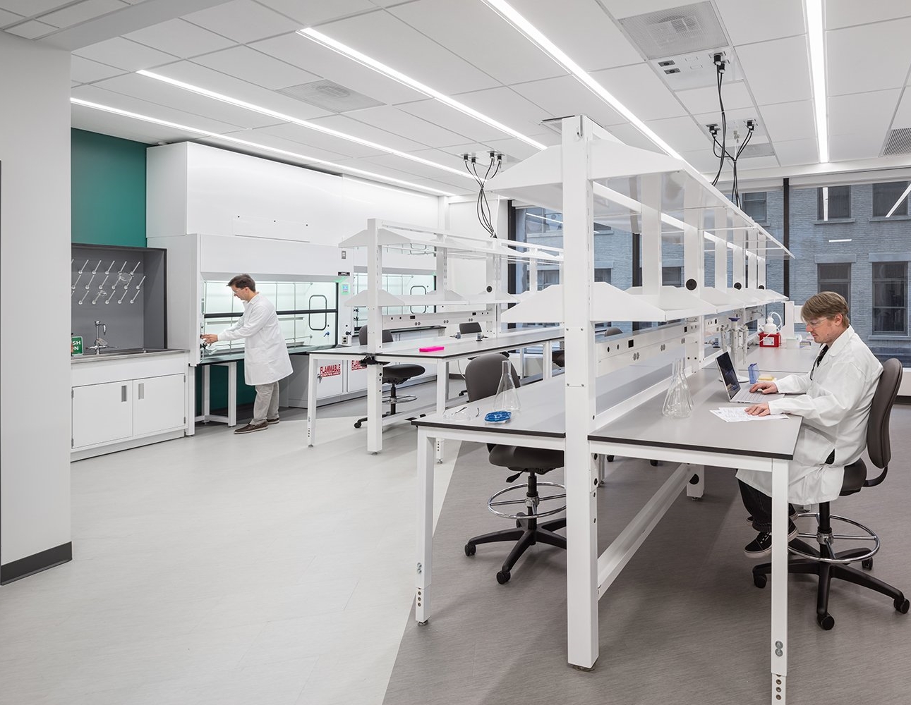 A modern laboratory with two scientists working. One scientist is standing and working with lab equipment near a fume hood, while the other is seated at a workbench using a laptop. The lab features clean, white countertops, overhead storage shelves, and bright overhead lighting, with large windows allowing natural light into the room.