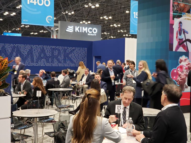 People seated at round tables in an exhibit hall. In the background is a large temporary blue wall